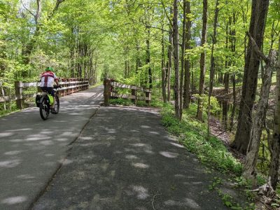 A paved trail with a wooden bridge extends through a deciduous forest with green spring leaves. A person with a white and red shirt and a green helmet is bicycling on the trail.