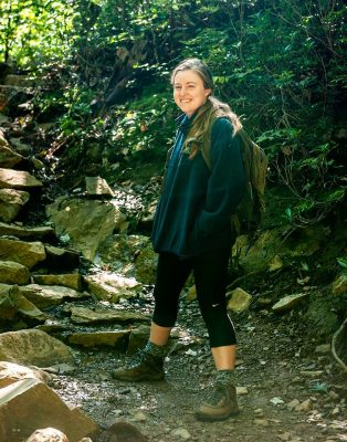 Laura stands smiling on a dirt trail at the base of a stone staircase. She has sandy hair and is wearing a teal sweatshirt and brown backpack.